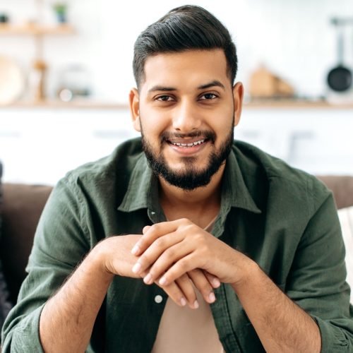 Portrait of confident attractive indian or arabian man, in casual stylish clothes, sitting at home in cozy living room on the background of the kitchen, looks at camera, smiling friendly